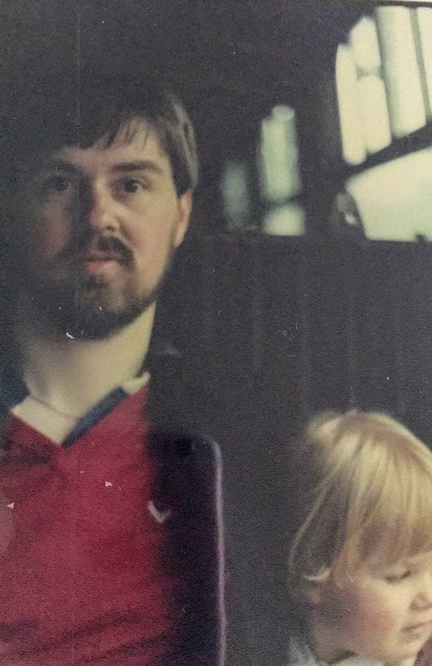 80s photograph of Sarah's dad wearing a red tshirt and jacket, looking young, with a beard. Sarah is 3 or 4 with very blonde hair, they are on a bus?