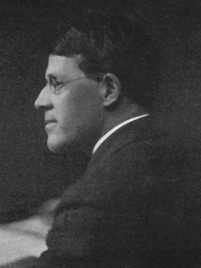 Appleby Matthews in a black and white photo, he is looking over his shoulder. He is wearing small classes and a suit jacket. 